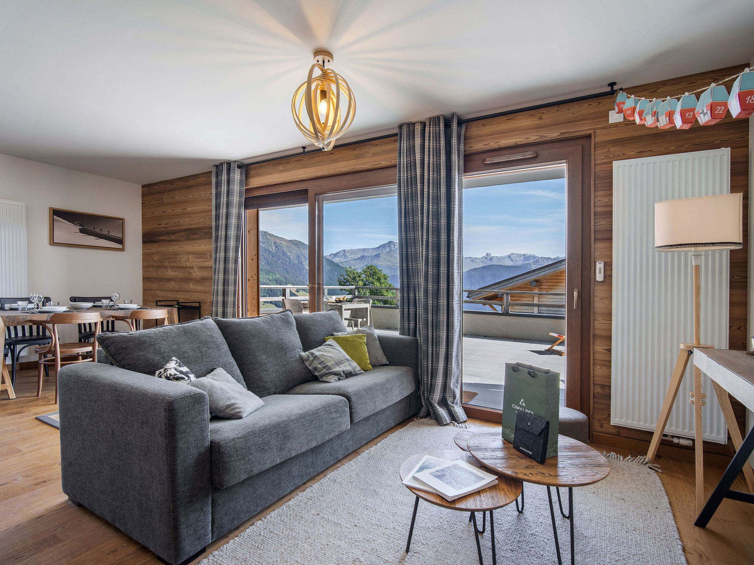 A modern living room with a grey sofa, wooden accents, a dining area with a table and chairs, and a view of mountains through large windows.