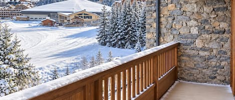 Un balcón de madera con un muro de piedra, con vistas a una pista de esquí nevada y edificios.