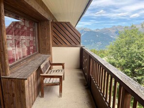 A wooden balcony with a bench, a window with patterned curtains, and a view of mountains.