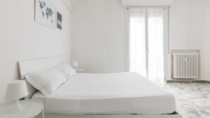 Een minimalistische witte slaapkamer met een bed, nachtkastjes, een radiator en een raam met dunne gordijnen.