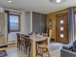 A dining area with a wooden table set for four, surrounded by chairs, in a room with wooden walls and a view of mountains through a window.