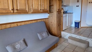 Ein kompakter Wohnraum mit einer Couch, einem kleinen Hocker, Holzschränken, einer Küchenzeile und einem Fliesenboden.