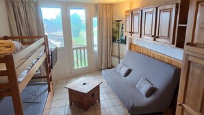Ein kleines Zimmer mit Etagenbett, einer Schlafcouch und einem kleinen Tisch. Große Fenster mit Vorhängen, ein Bild an der Wand und Holzschränke über der Couch.