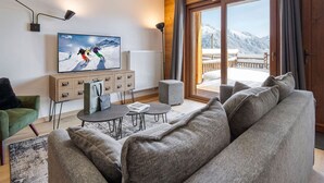 A modern living room with a large TV, a sofa, a coffee table, and a view of snowy mountains through a glass door. - Comfortable T2 cabin for 6, ski shuttle, parking included (Les Saisies)