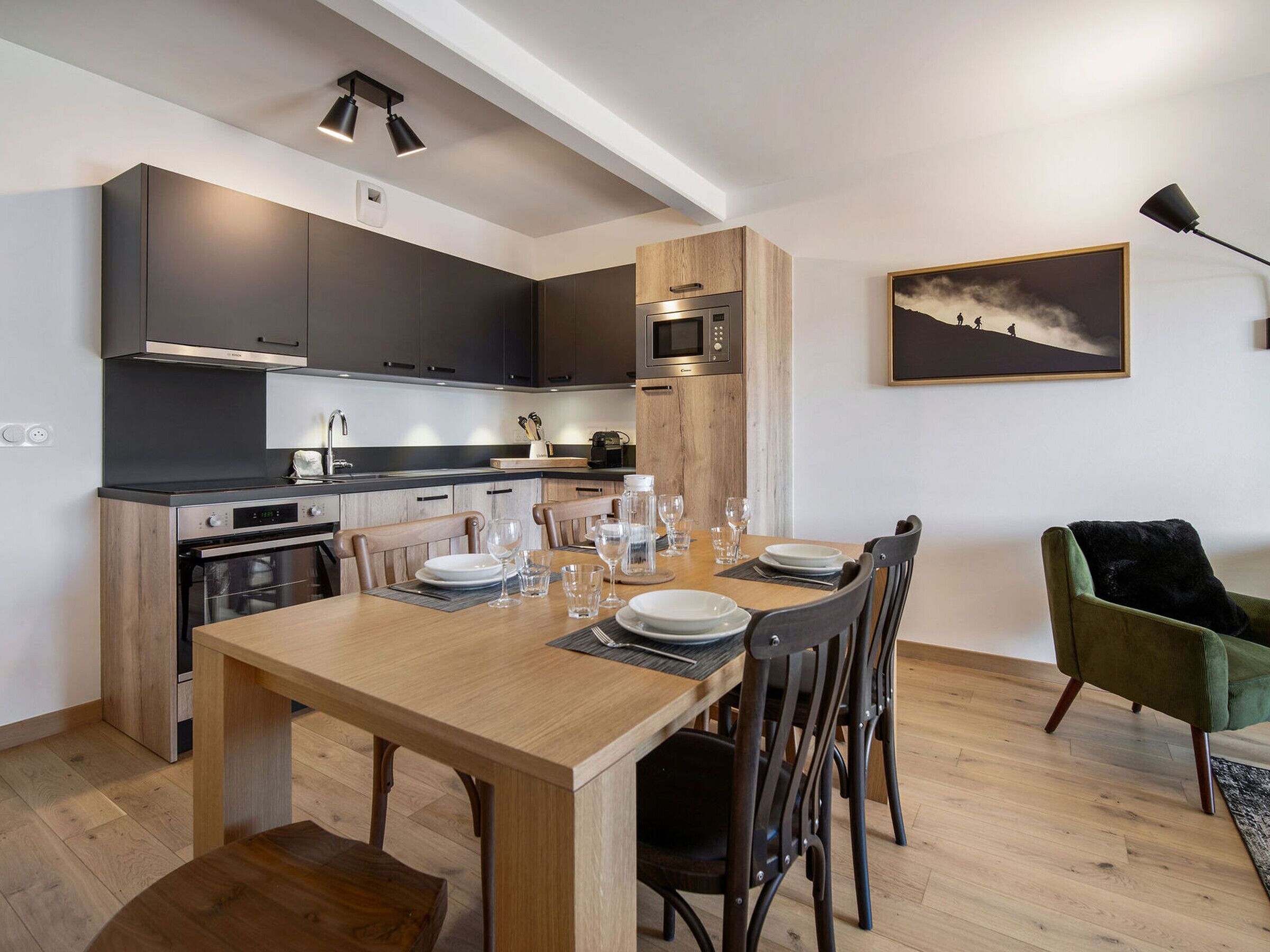 A modern kitchen with dark cabinets, a wooden dining table set for four, and a green armchair.