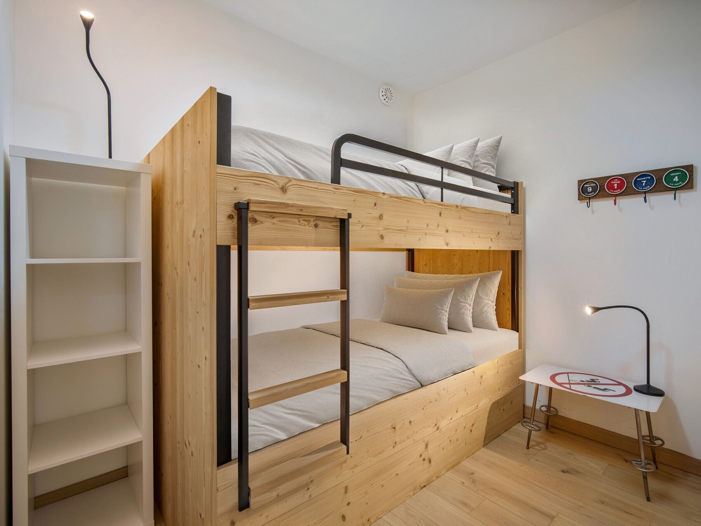 Een stapelbed met een houten ladder, een klein tafeltje met een lamp en een wandplank met een bordje.