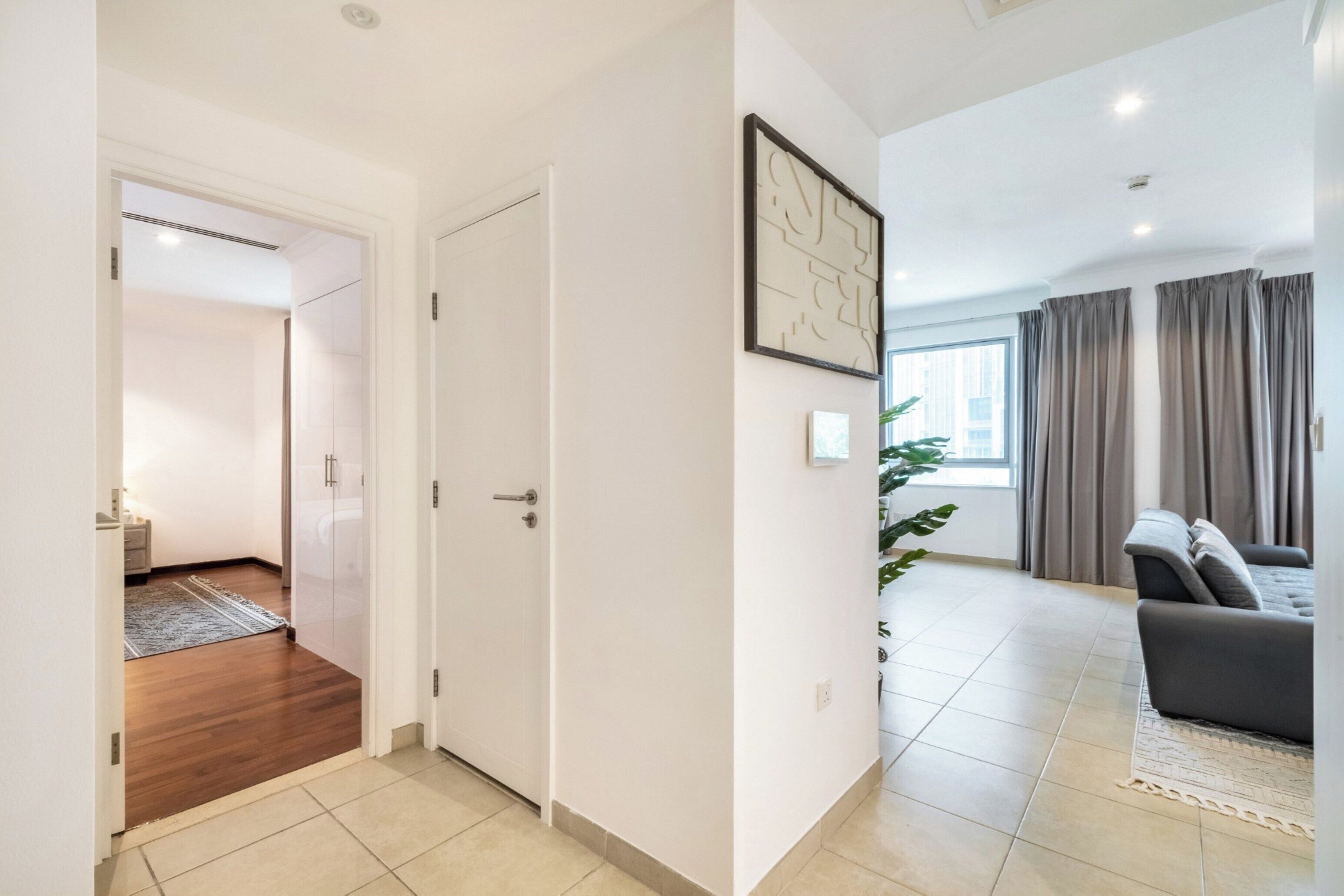 A modern apartment with a wooden floor hallway, a living area with a grey sofa, and a large window with curtains.