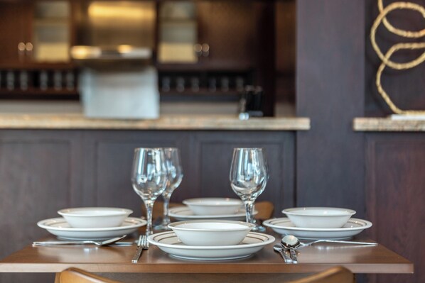 Una mesa de comedor dispuesta con platos, tazones y copas de vino, con un fondo de madera oscura y una cocina moderna al fondo.
