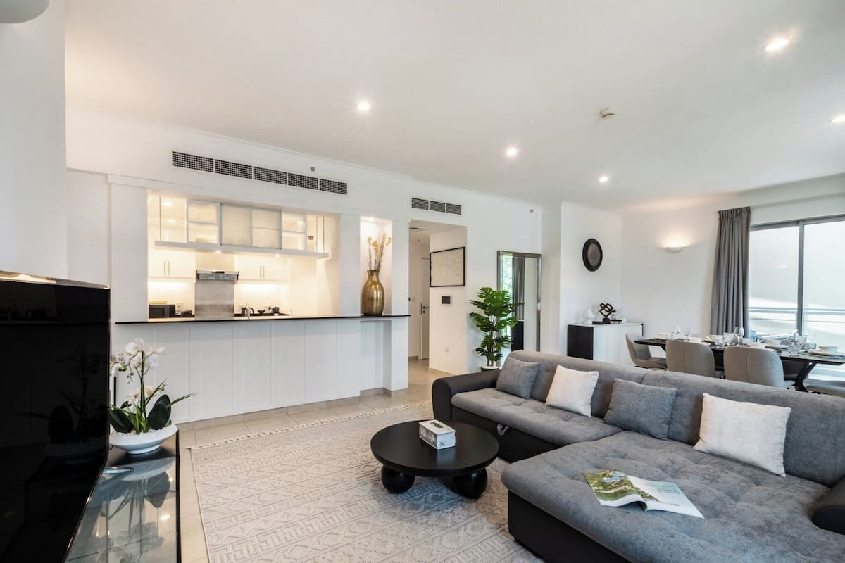 A modern living room with a grey sofa, a coffee table, and a TV. There is a kitchen area with white cabinets and a dining area with a table and chairs.