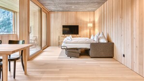 Een moderne kamer met houten inrichting, een bed met kussens, een televisie en een zithoek met stoelen.