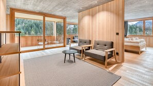 Een modern interieur met een houten vloer, een bank, een salontafel en een bed. Grote ramen bieden uitzicht op een bebost gebied.