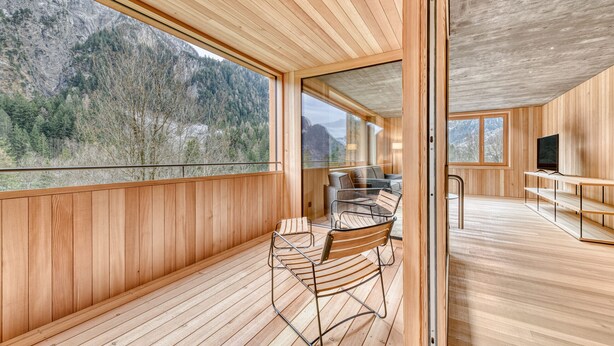 Een modern interieur met houten lambrisering, een glazen deur naar een balkon met uitzicht op de bergen, en een zithoek met metalen stoelen en een tafel.