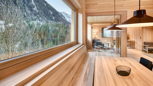 Un interior moderno con paredes y suelos de madera, un ventanal con vistas a la montaña, y un comedor con mesa y sillas.