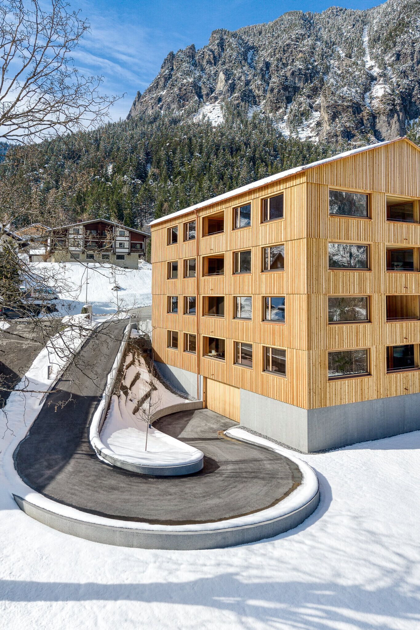 A modern building with multiple windows, a curved driveway, and a snowy mountain backdrop.