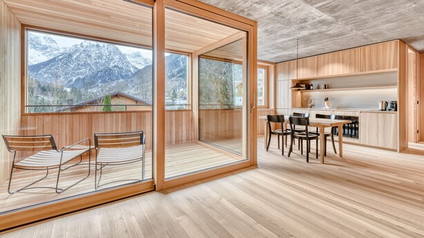 A modern kitchen with wooden cabinets, a dining area with chairs, and large windows offering a mountain view. - Appartmentshaus Studa 29 - Studa 7 (Brand)
