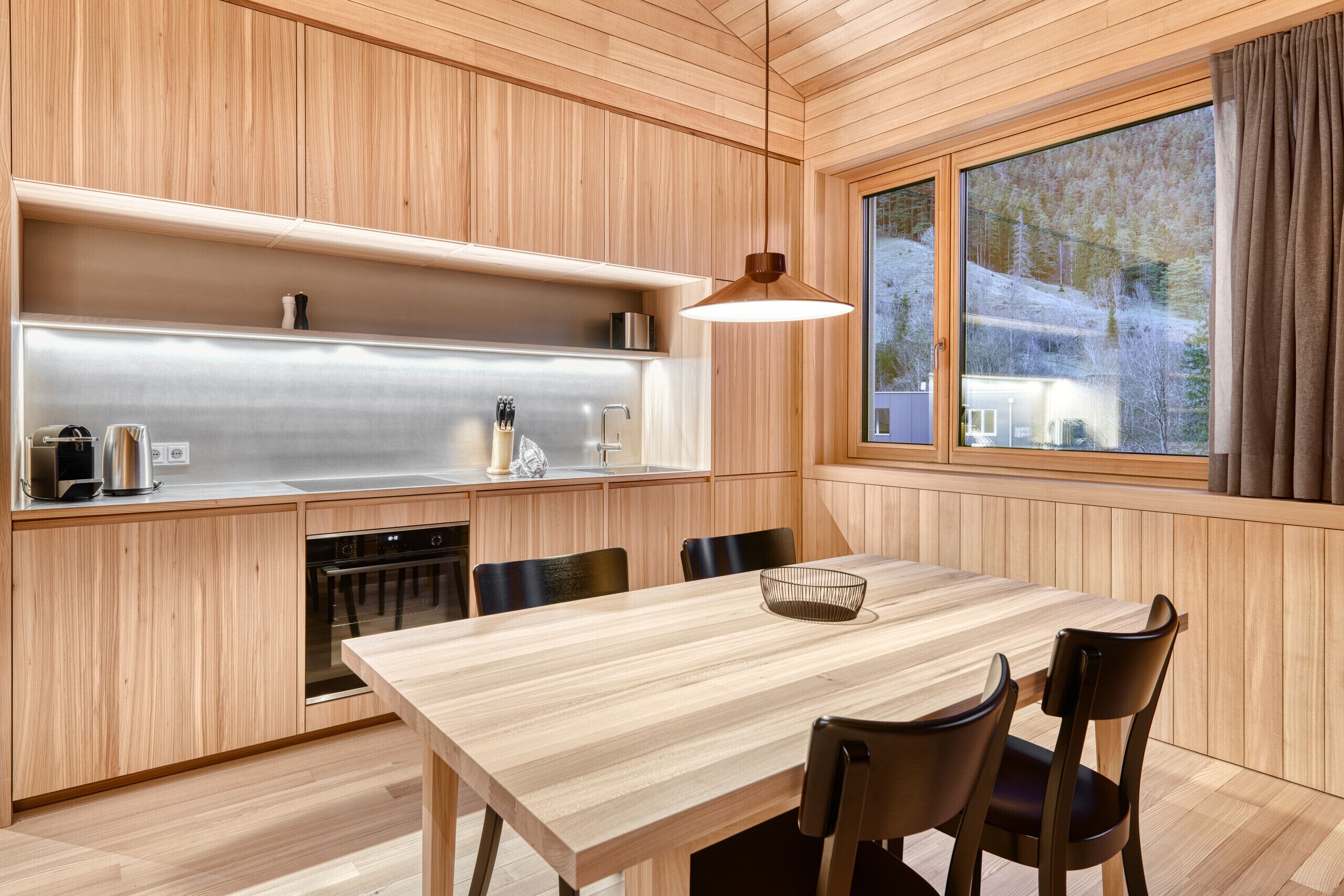 Une cuisine moderne avec des meubles en bois, des appareils électroménagers en acier inoxydable, une table à manger avec des chaises et une grande fenêtre donnant sur un paysage enneigé.