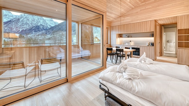 A modern hotel room with a large sliding glass door leading to a balcony, a separate dining area, and a cozy bed.