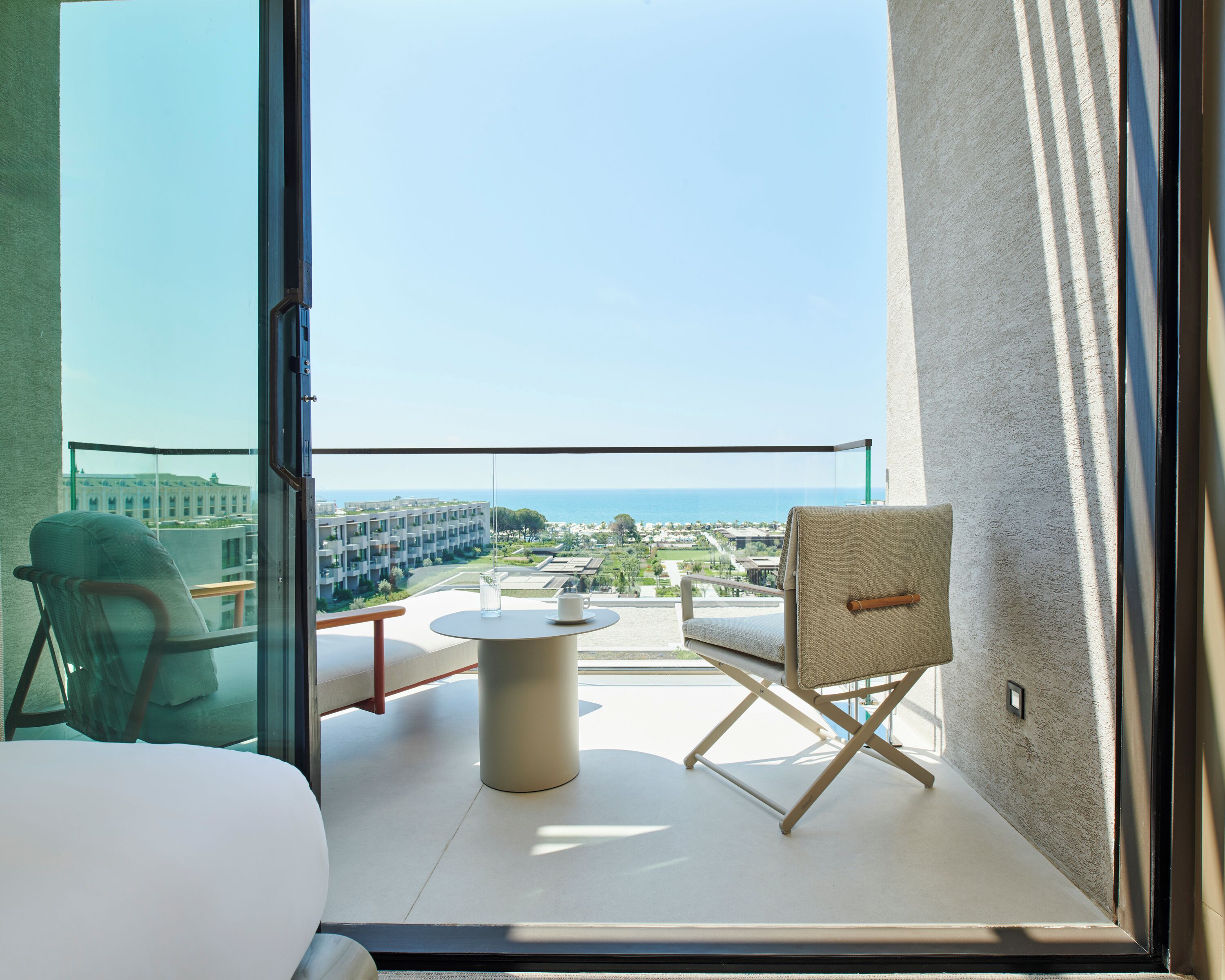 Ein Balkon mit Glastrennwand, zwei Stühle, ein runder Tisch und Blick auf eine Küstenlandschaft mit Gebäuden.
