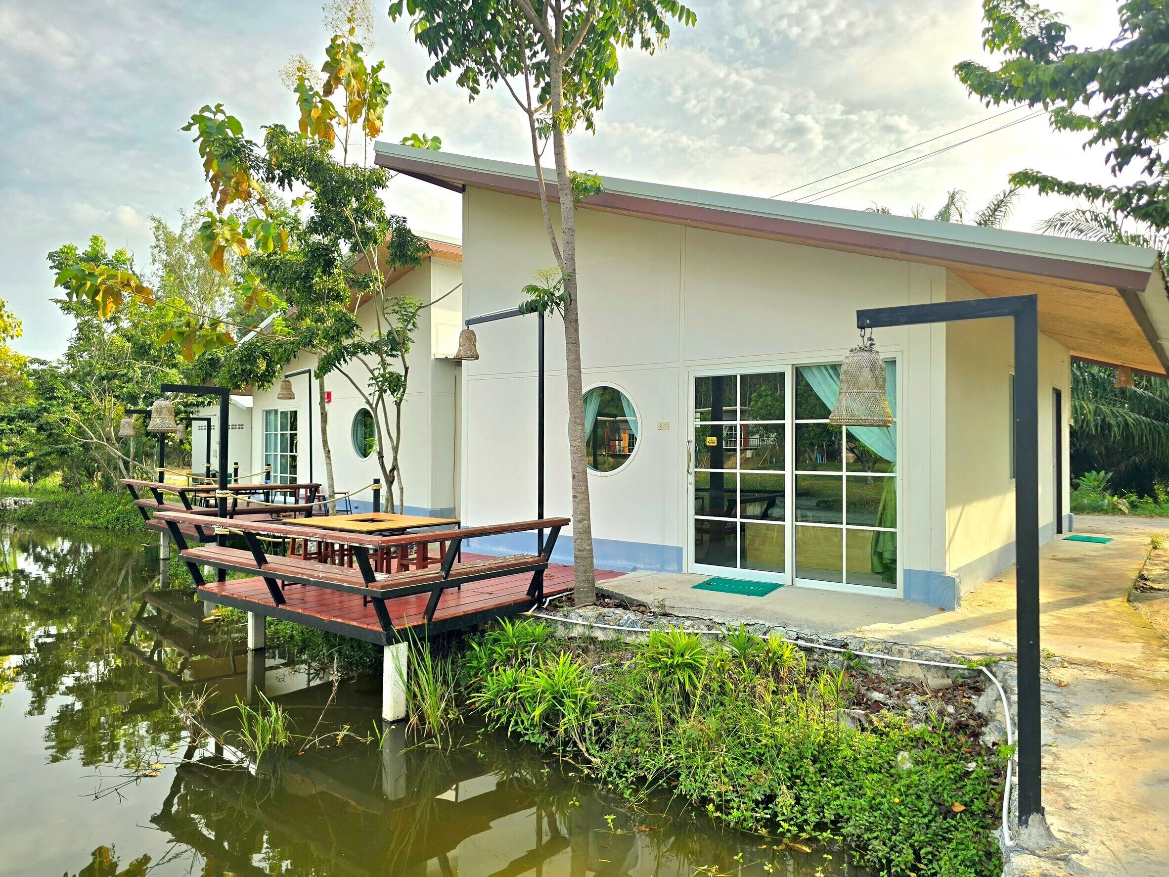 Een modern gebouw met een overdekt terras aan een rustig water, omgeven door weelderige begroeiing.