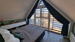 A bedroom with a sloped ceiling, a bed with a quilted mattress, a nightstand with bottles, and a window with curtains.
