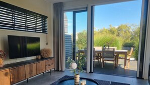 Una sala de estar moderna con un televisor de pantalla plana, una mesa de centro redonda con objetos decorativos, una alfombra oscura y un ventanal corredero que da a un patio exterior con mesa y sillas de madera.