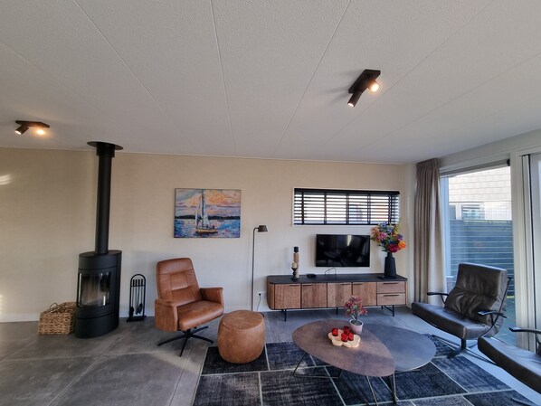 A modern living room with a fireplace, leather chairs, a coffee table, a TV, and a window with blinds.