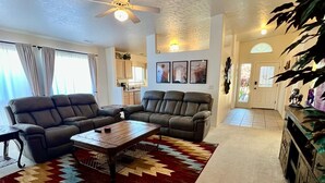 A spacious living room with a large sectional sofa, a coffee table, and a patterned rug. There is a ceiling fan, a dining area in the background, and a kitchen visible through an open door. - Large Prescott Home w/3-car Garage Central & Quiet (Prescott)