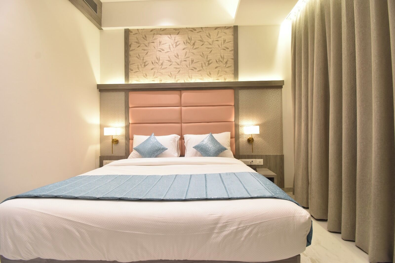 Camera d'albergo con un letto grande, due lampade ai comodini, una testiera con motivo a foglia e una finestra con tende.