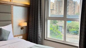 Ett hotellrum med en stor säng, en sänglampa och utsikt över byggnader och grönområden utanför.