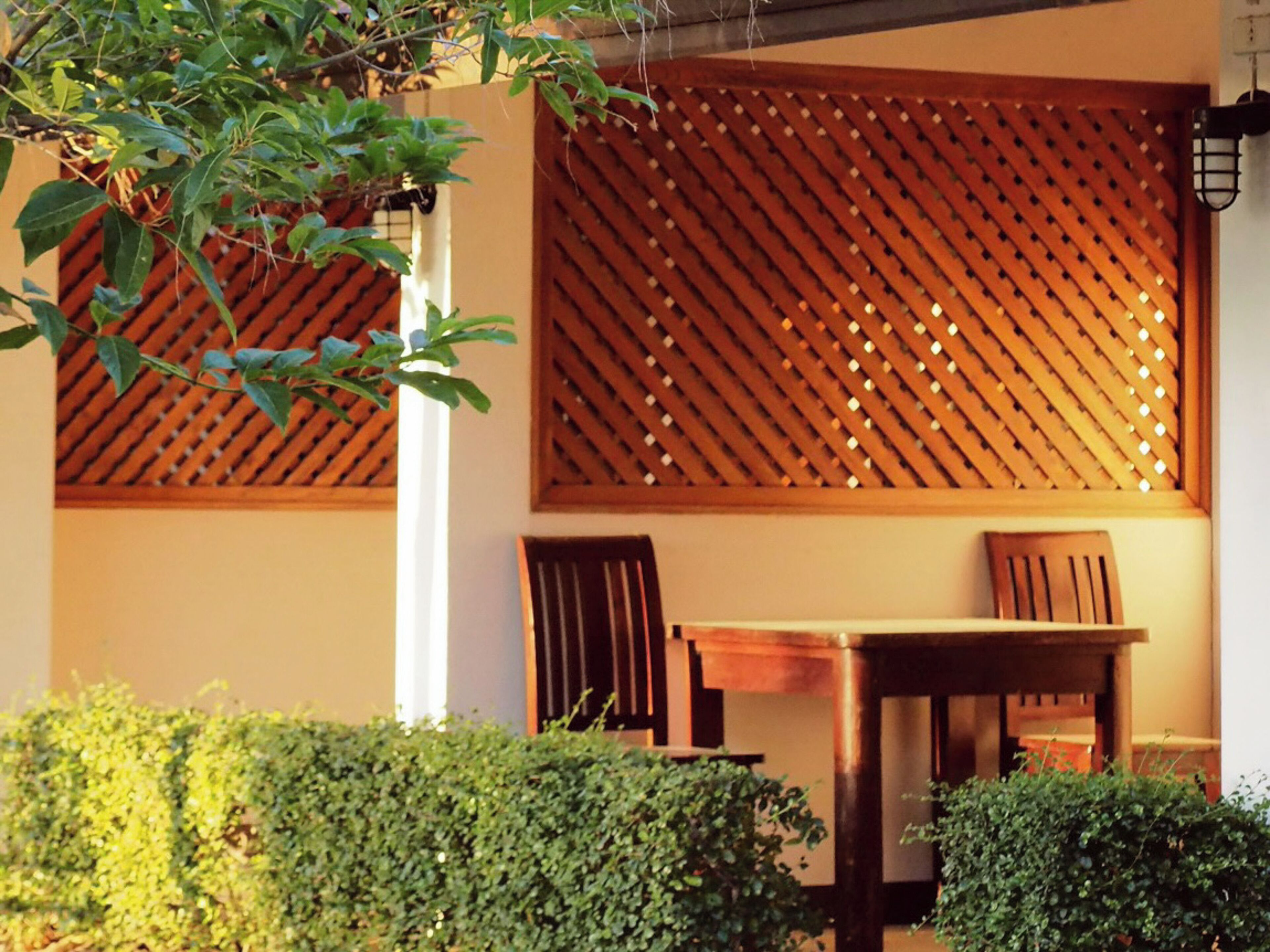 A wooden table and chairs set against a wall with a lattice design, surrounded by greenery.