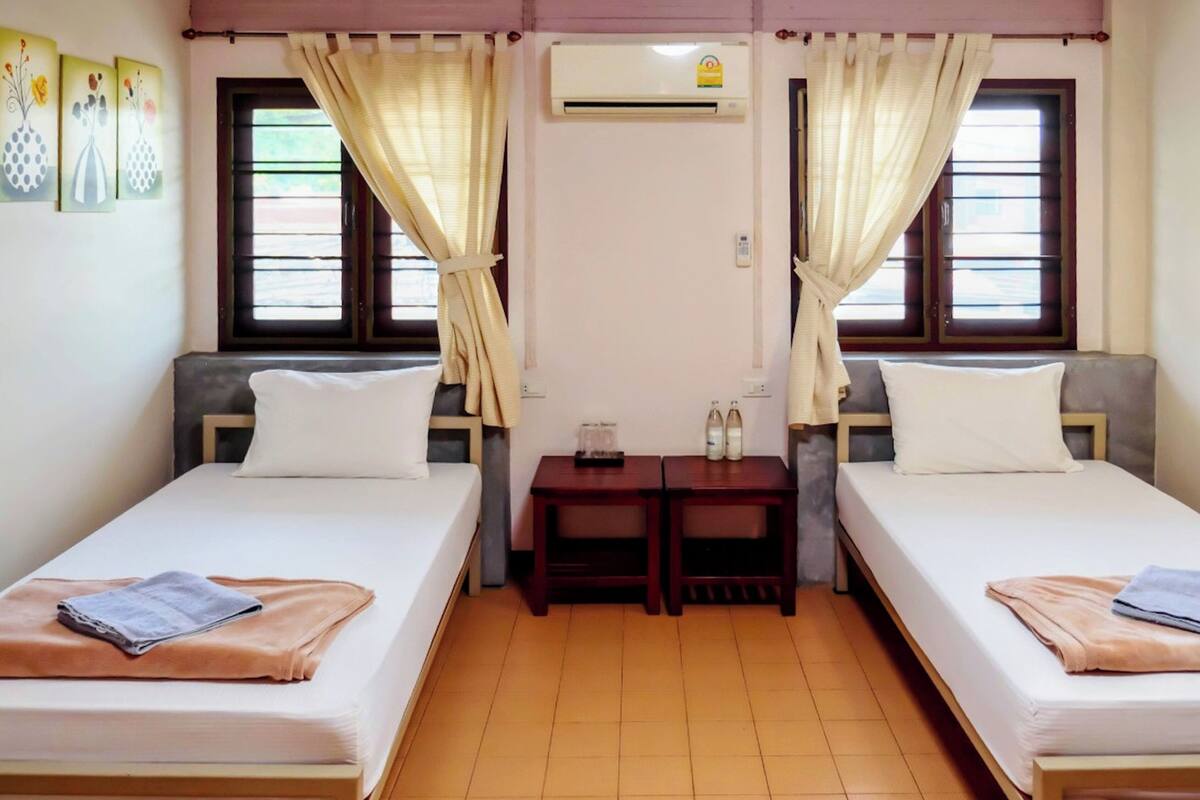 A room with two single beds, a small table with bottles, and an air conditioning unit.