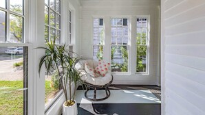 Une pièce aux murs blancs et dotée d’une grande fenêtre, avec un fauteuil à bascule muni d’un coussin et une plante en pot.