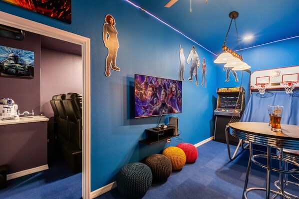 A room with a blue wall, a TV mounted on it, a basketball arcade game, and colorful ottomans.
