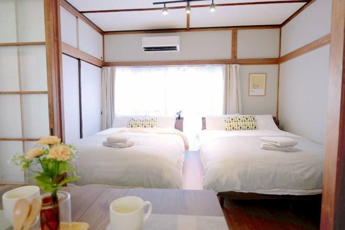 A hotel room with two beds, a window with curtains, a small table with a vase of flowers, and an air conditioning unit.