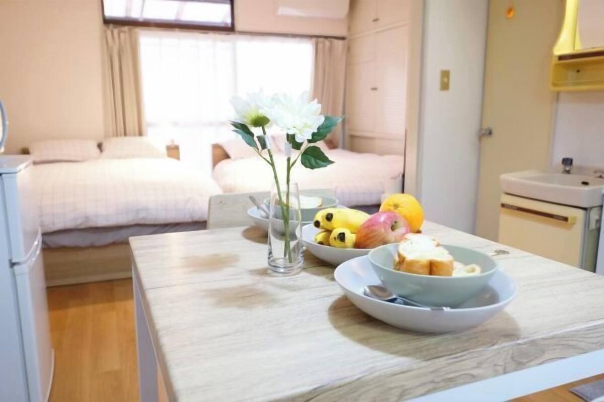 A bedroom with a bed, a bedside table with a vase, and a breakfast setup with fruits and bread.
