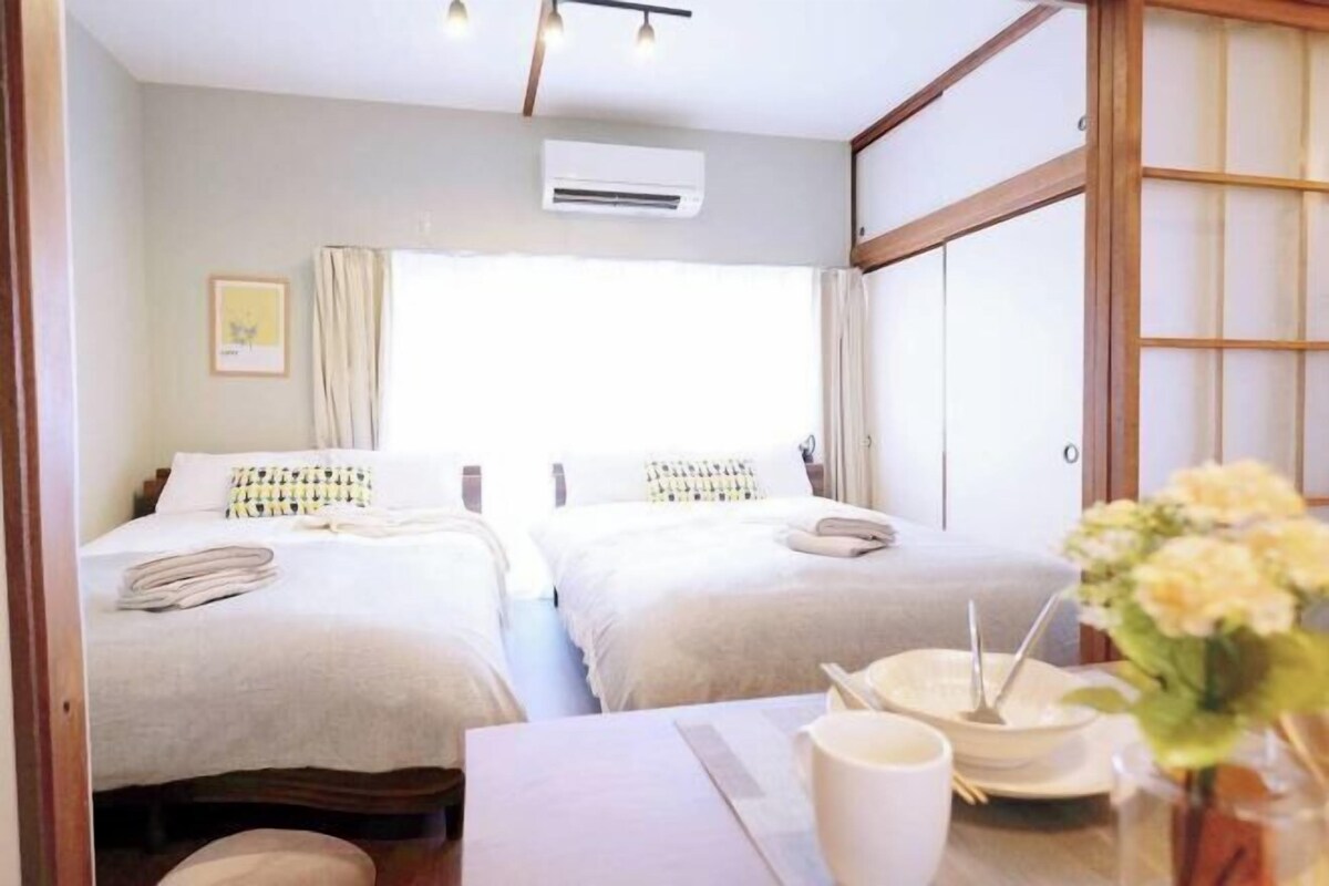 A hotel room with two beds, a dining table with a vase of flowers, and an air conditioning unit on the wall.