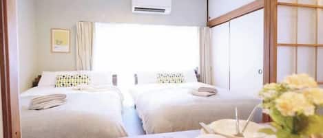 A hotel room with two beds, a dining table with a vase of flowers, and an air conditioning unit on the wall.