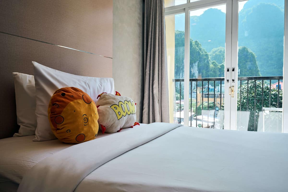 A hotel room with a bed, pillows, a window with a view of mountains, and a balcony.