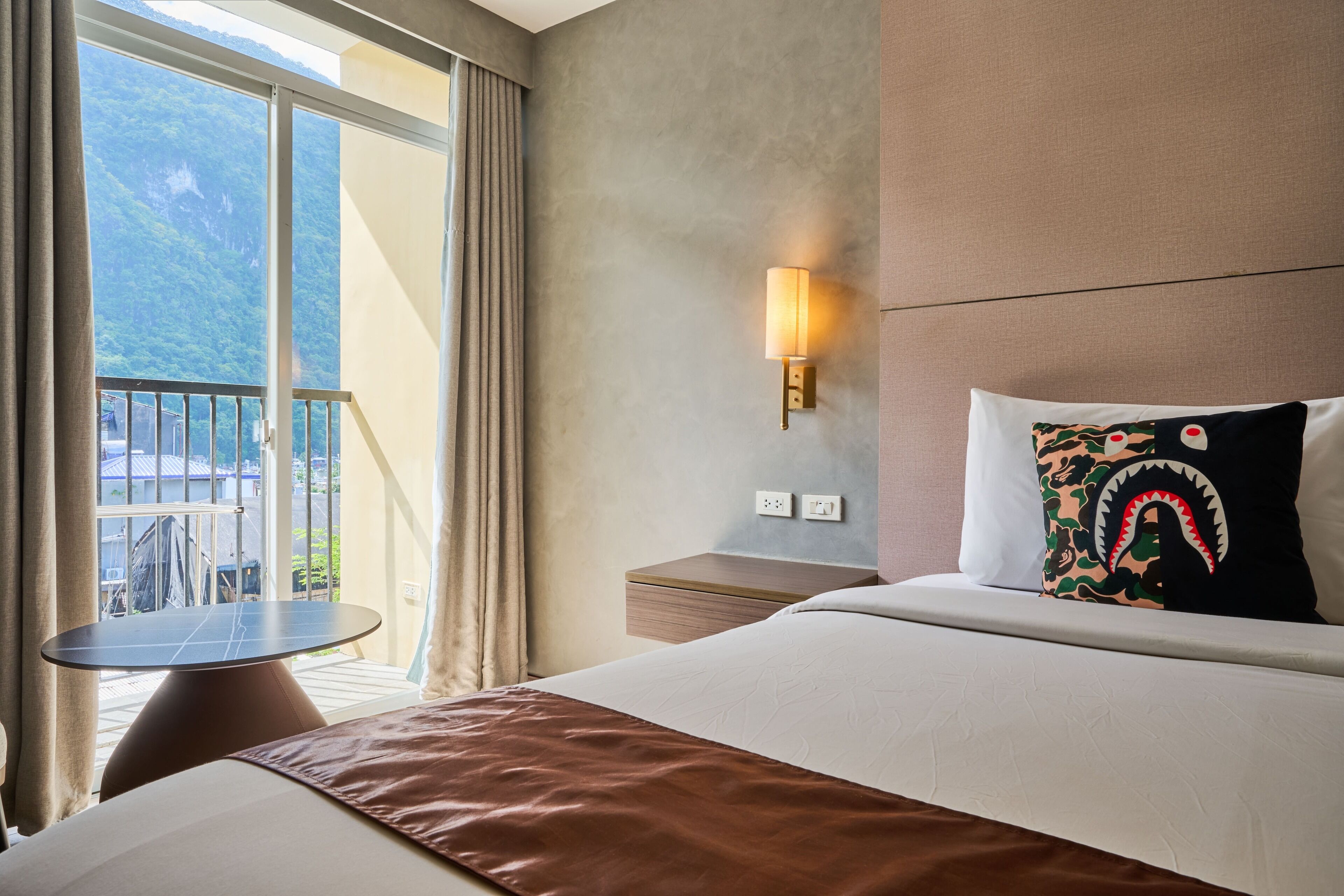 A hotel room with a bed, a bedside table, a lamp, a view of a balcony, and a window.