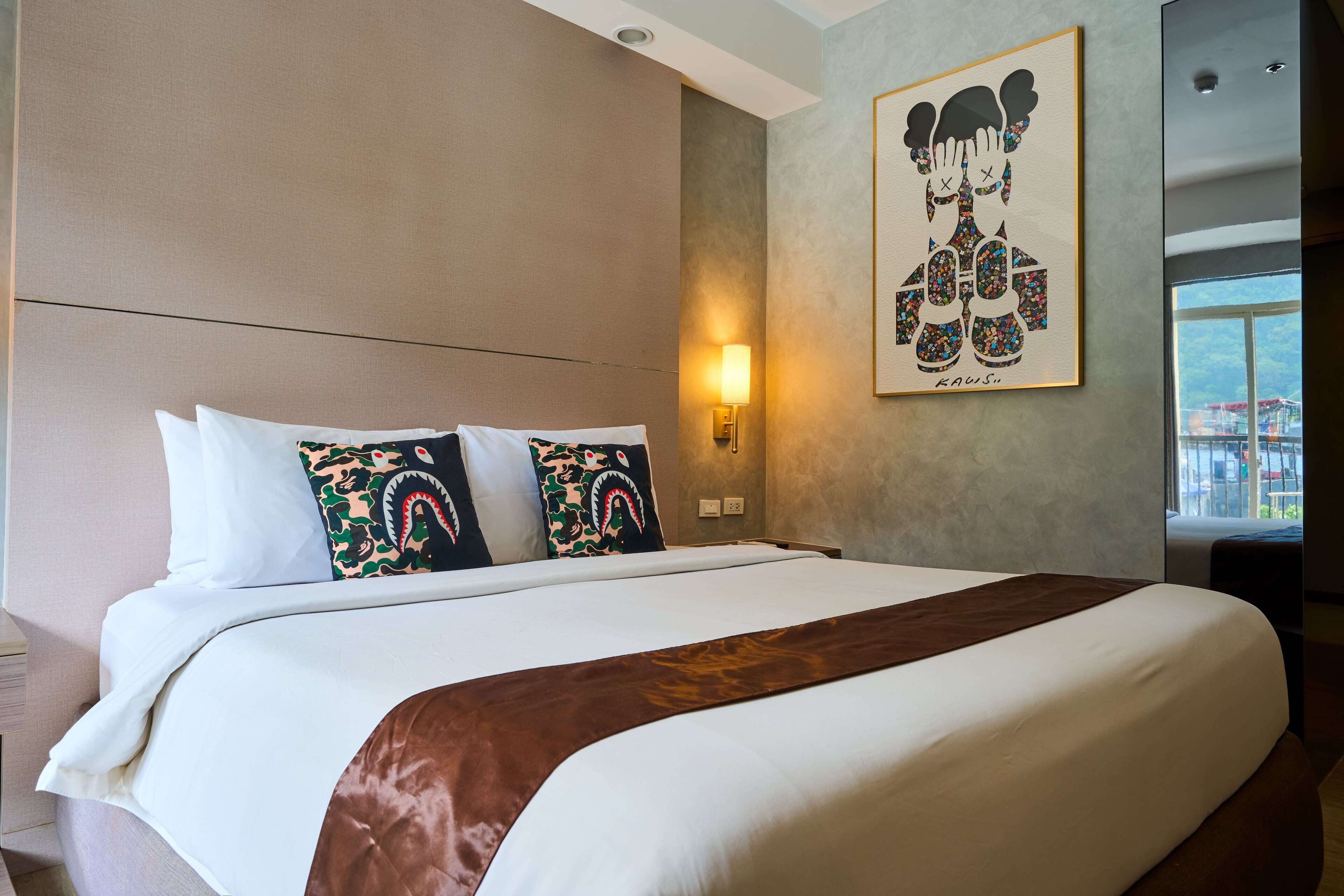 A hotel room with a large bed, a wall-mounted artwork, a bedside lamp, and a window with a view.