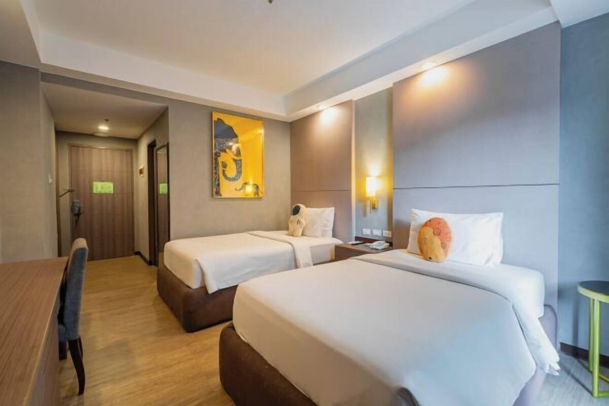 A hotel room with two beds, a desk, a chair, a mirror, and a wall art piece.