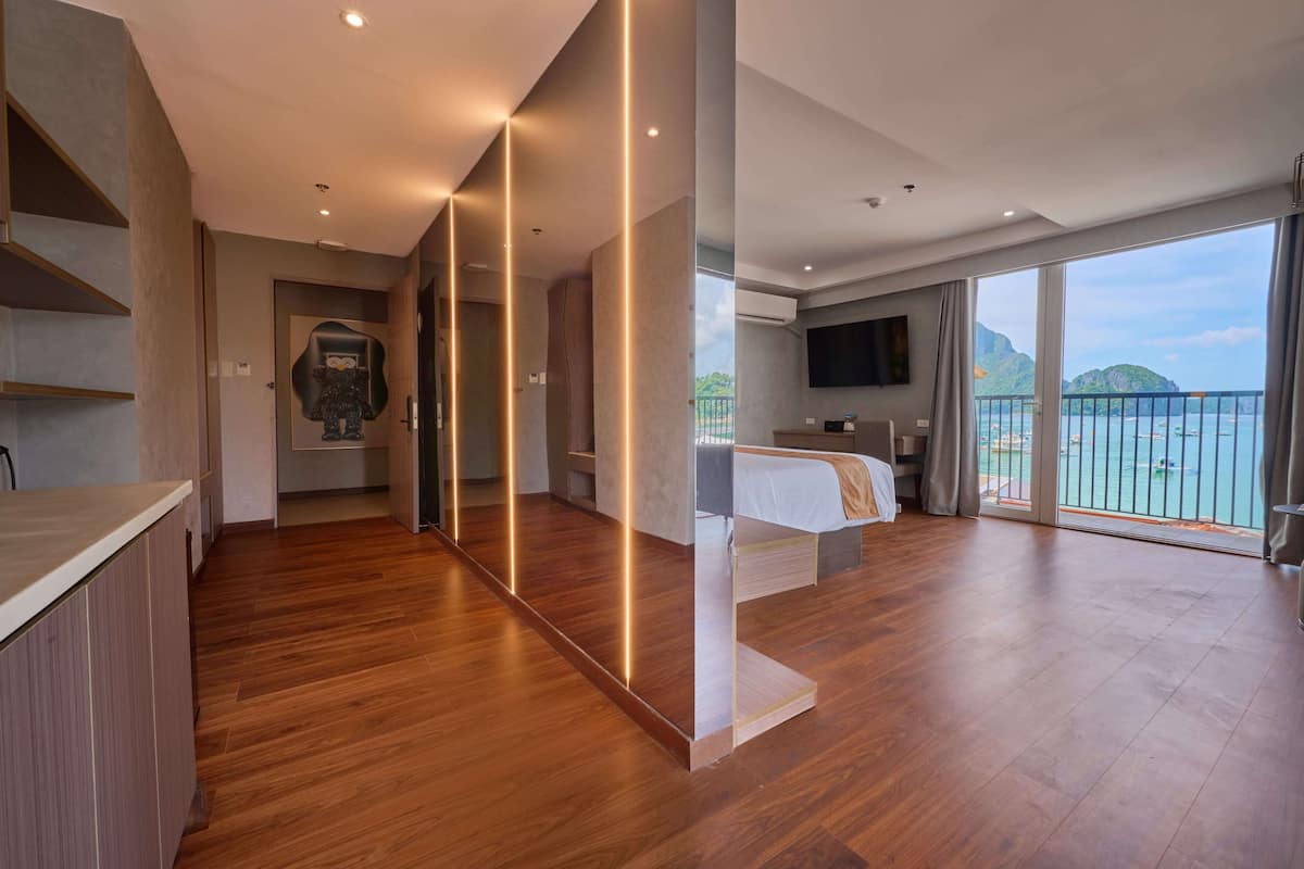 A modern hotel room with a wooden floor, a bed, a TV, and a balcony with a view of the sea.