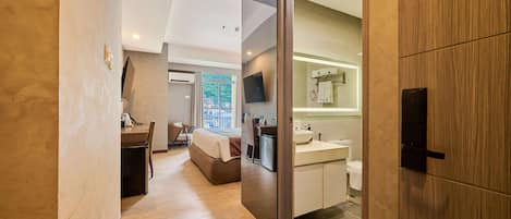 A modern hotel room with a wooden floor, a bed, a desk with a chair, a bathroom with a sink and mirror, and a television mounted on the wall.