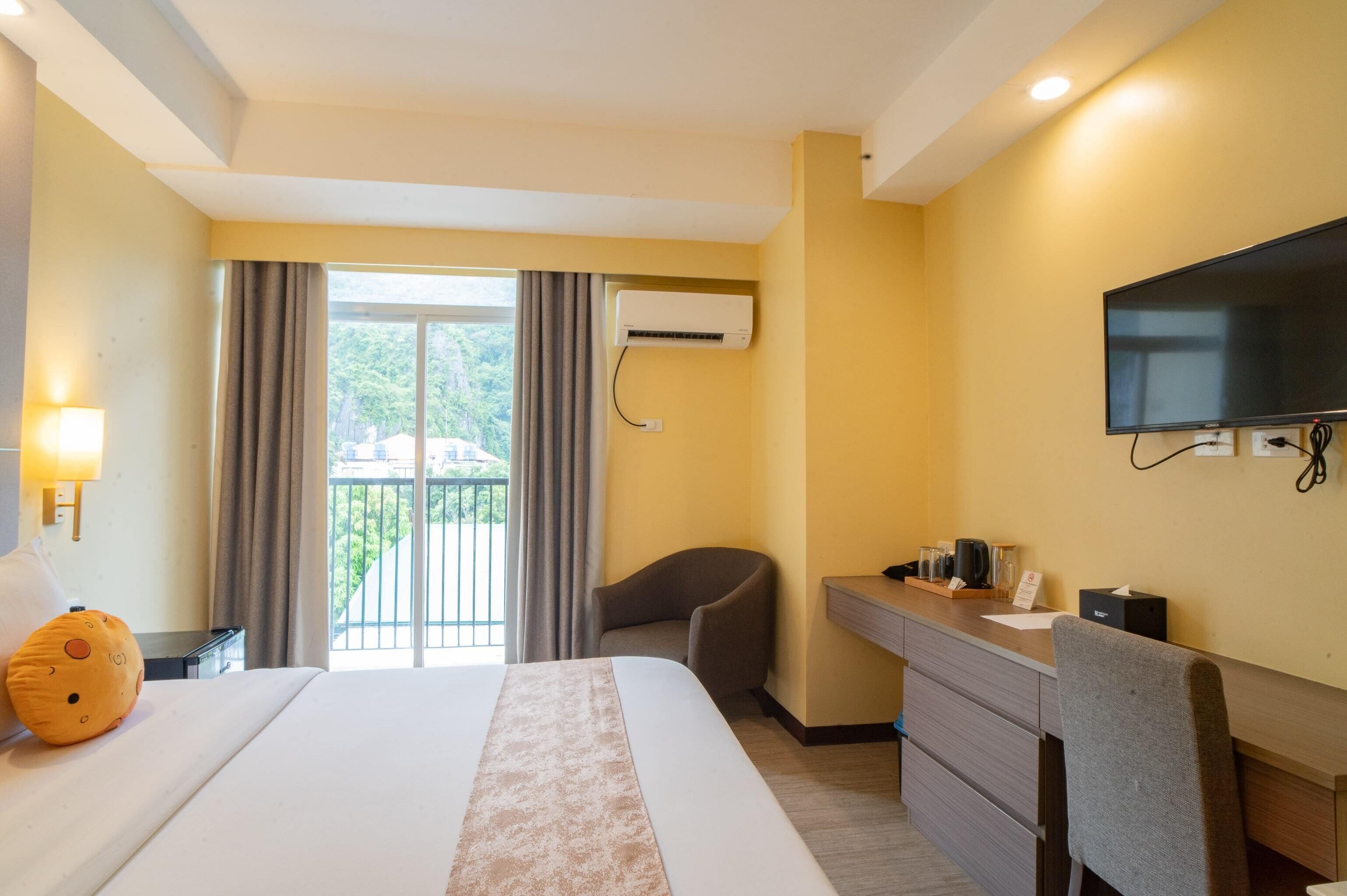 A hotel room with a bed, a desk, a chair, a TV, and an air conditioning unit.