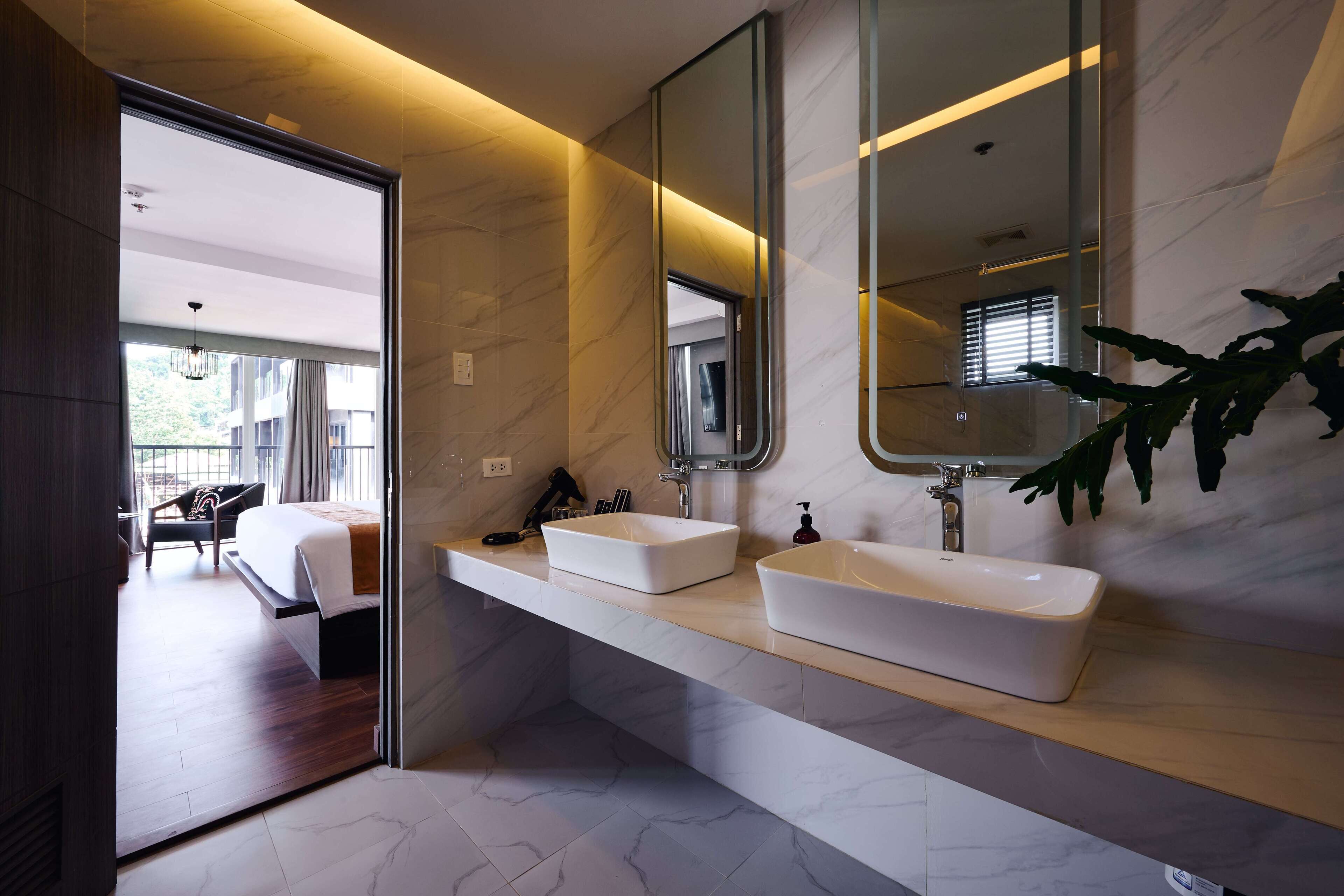 Une salle de bain moderne avec deux lavabos, un grand miroir et une vue sur la chambre.