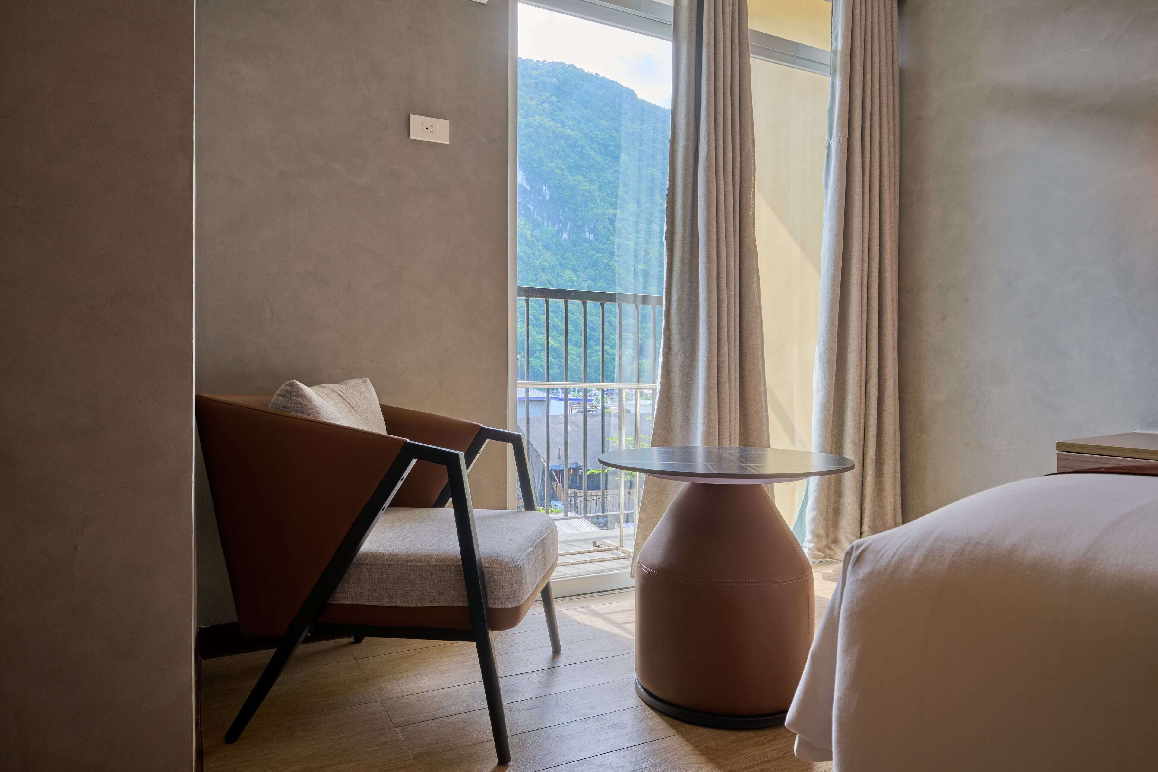 A hotel room with a bed, a chair, a small table, and a balcony with a view of a mountain.
