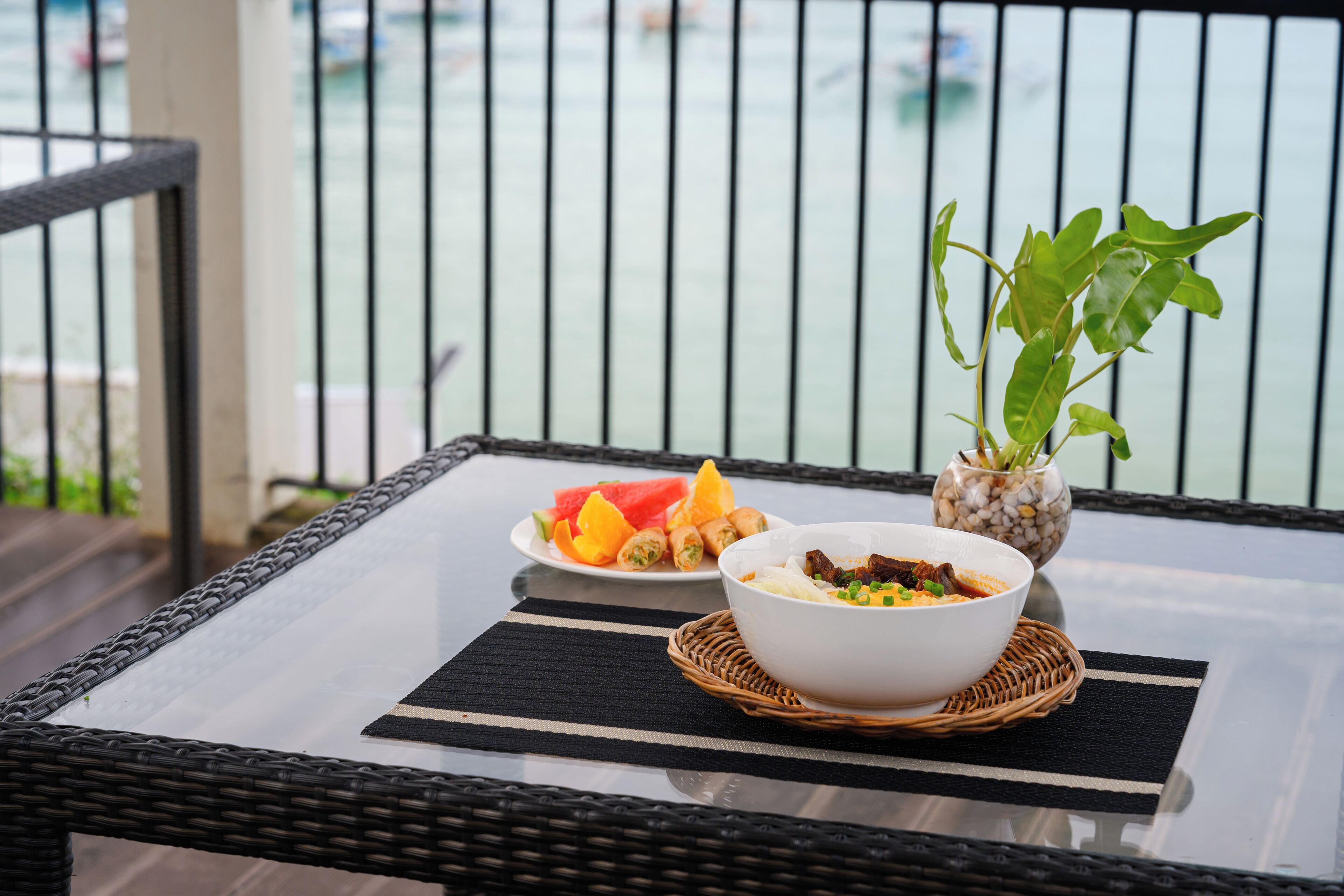 Una mesa de comedor con un bol de comida, un plato de fruta y una planta en maceta en un balcón con barandilla y vistas al mar.