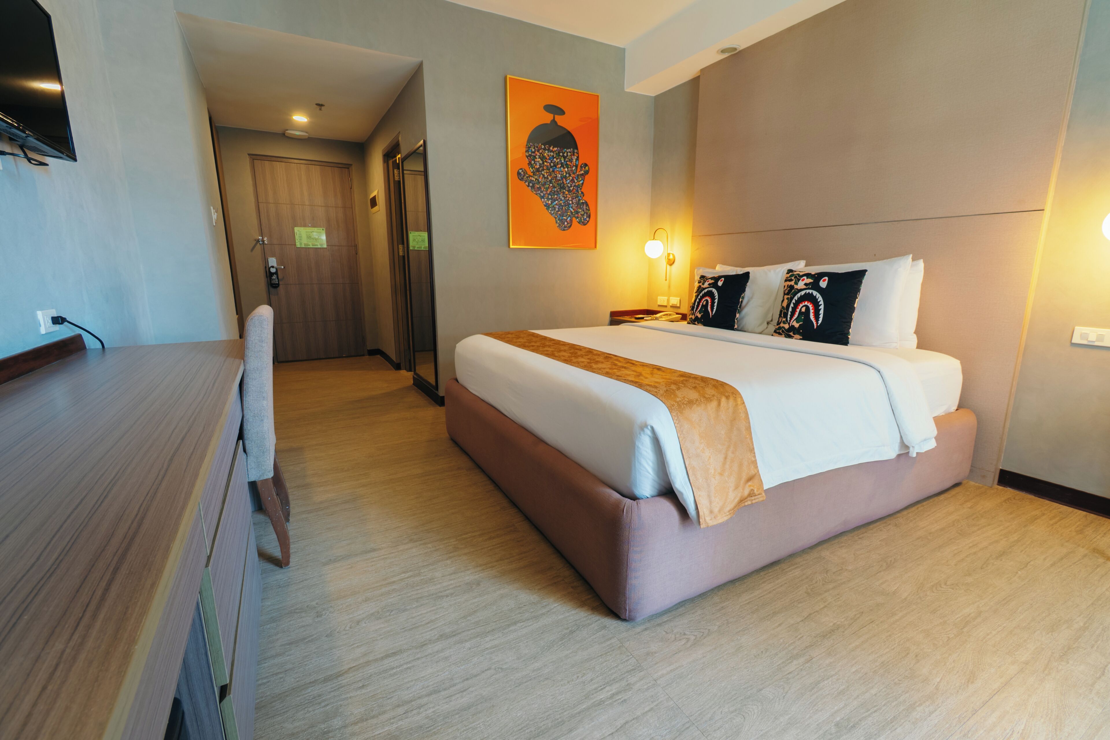 A hotel room with a bed, a desk, a chair, a door, and a wall-mounted artwork.
