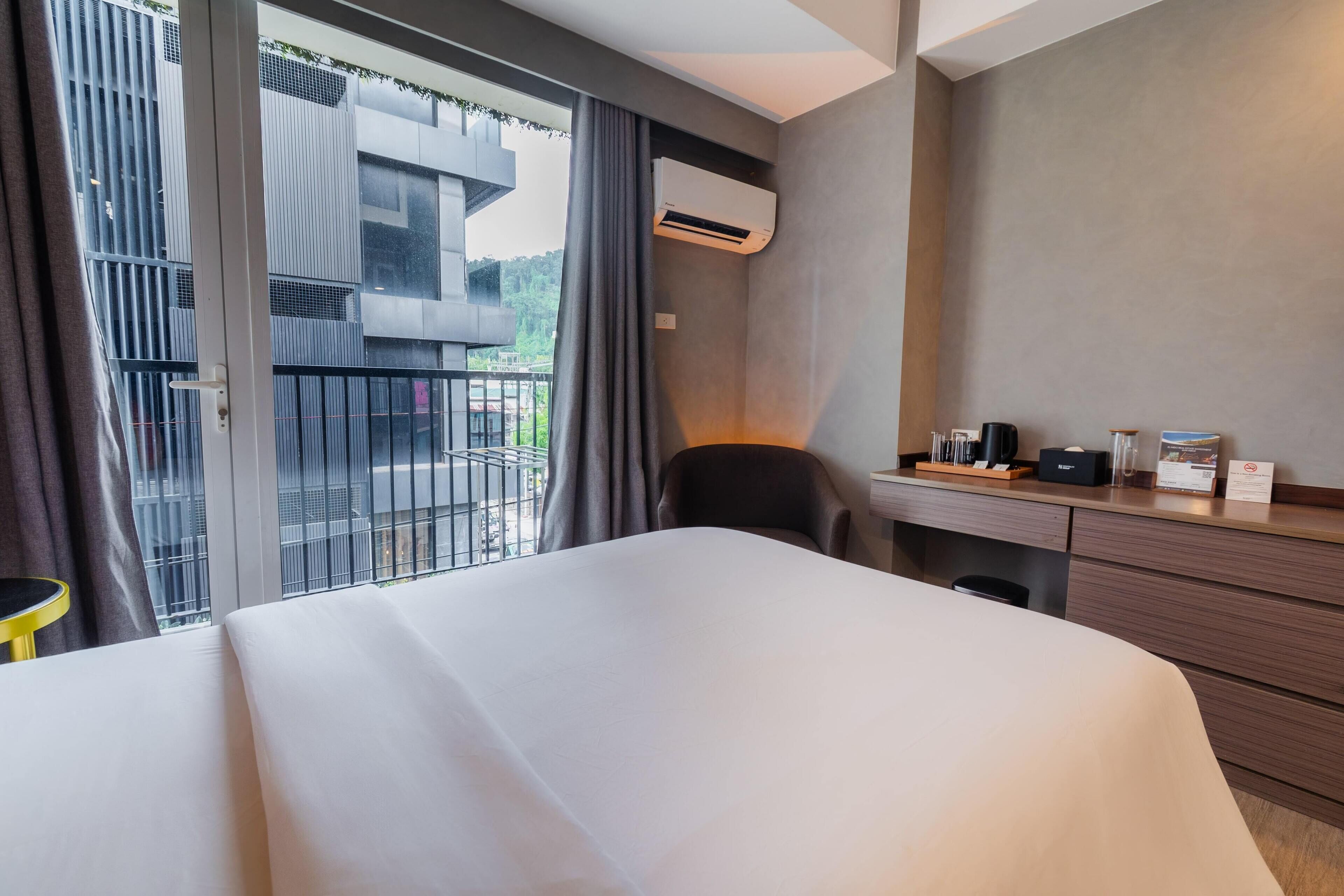 A hotel room with a bed, a desk, a chair, an air conditioning unit, and a view of a balcony with railings.