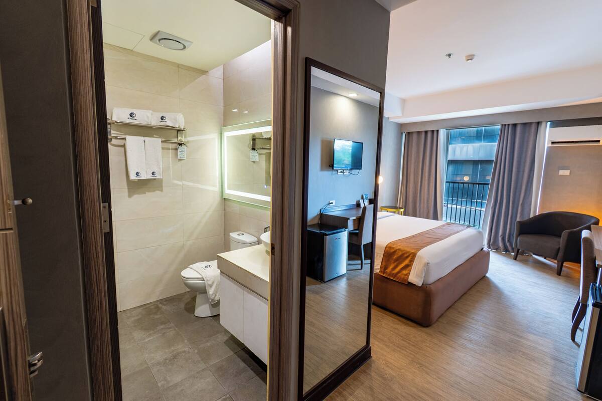 A hotel room with a bed, a desk, a chair, a bathroom with a toilet and sink, and a large mirror.